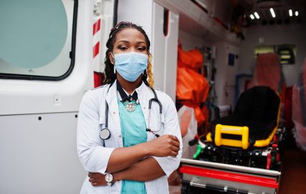African american female paramedic in face protective medical mask standing in front of ambulance car.